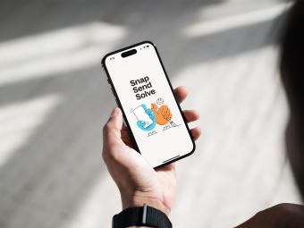 Hand holding a smartphone displaying the Snap Send Solve app screen with blue and orange illustrated characters and bold text reading “Snap Send Solve”.