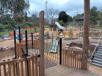 A modern playground with wooden structures with multiple slides in a bushy park setting.