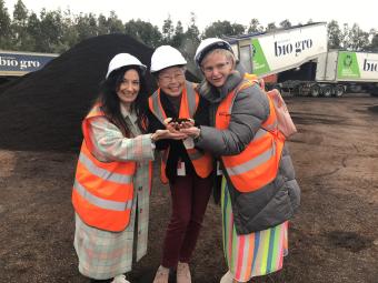 Three Manningham councillors smiling and wearing hard hats, stand holding a handful of compost. In the background is a large pile of compost and a Bio Gro truck.