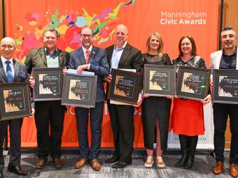 Five men and two women stand in front of an orange banner with the text Manningham Civic Awards in the top right. They are smiling and holding their awards in front of them.