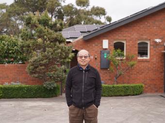 A man standing in front of a brick house with solar panels on the roof, surrounded by greenery.