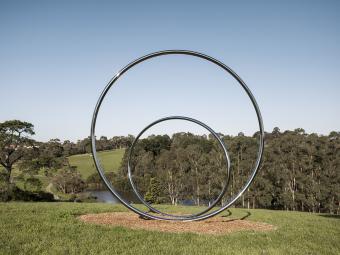 Two large polished stainless steel circles sit on a hill overlooking an open grassy expanse with gum trees and a lake in the background.