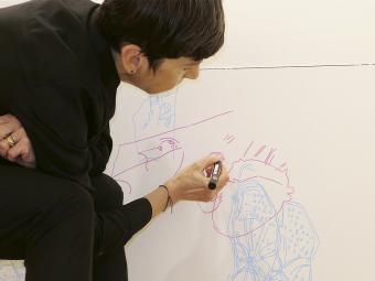 Woman dressed in black sits on stool and draws a face onto the wall, child's drawing with the word Max is stuck to the wall to her right