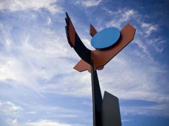 Large steel sculpture comprising bold geometric shapes in black, brown and blue sits against a blue sky.