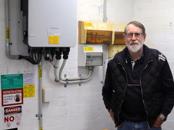 A man wearing glasses and a black jacket stands with his hands in his pockets. He is in a garage and a solar battery and a switchboard can be seen on the wall to his right.