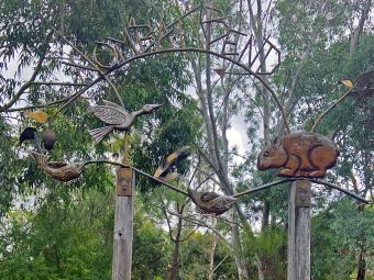 A concrete path leads up to two wooden poles with a gate between them. Above the poles sits a twisted metal sign with the words 'Wombat Bend'. A bushy parkland is in the background.