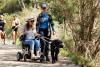 A woman in a wheelchair holds the lead of a black labradoor as another woman walk by her side. They are on a dirt path in a bush setting with other walkers and dogs behind them.