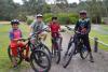 Four kids and an adult smile  to the camera, posing with their mountain bikes on a dirt path surrounded by greenery.