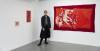 Artist Shannon Slee, wearing a black skirt and denim jacket, stands in the corner of a room next to a large red tapestry which is hanging on the while walls on the right, and a small framed red tapestry on the left.