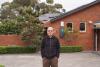 A man standing in front of a brick house with solar panels on the roof, surrounded by greenery.
